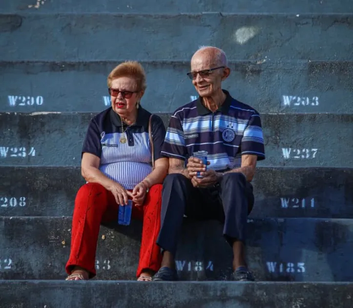 Casal de idosos, sentados lado a lado, vestindo a camisa do ASA, no Coaracy da Mata Fonseca